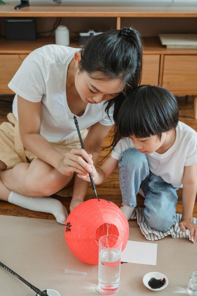 An Asian mother guides her son in painting a bright red lantern indoors, embracing cultural traditions.